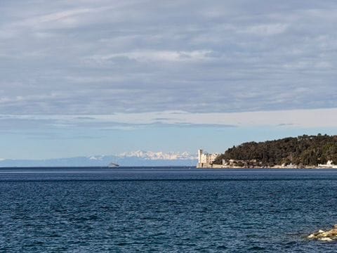 Blick über das Meer auf Schloss Miramare an der bewaldeten Küste, dahinter verschneite Berge